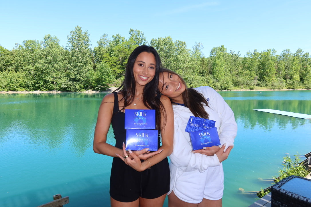 Two women holding blue Skilk Skin boxes by a lake with trees in the background